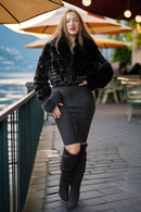A woman stands on an outdoor patio by a railing, wearing a black faux-fur cropped jacket, a fitted black mini skirt with a front cutout, and black over-the-knee boots.