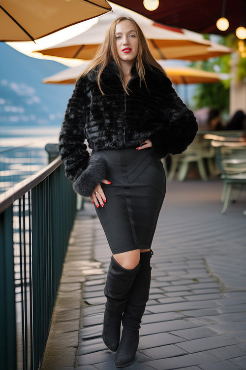 A woman stands on an outdoor patio by a railing, wearing a black faux-fur cropped jacket, a fitted black mini skirt with a front cutout, and black over-the-knee boots.
