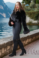 A woman posing outdoors by a lake wearing a black faux fur cropped jacket with matching cuffs, a dark gray pencil skirt, and black over-the-knee boots.