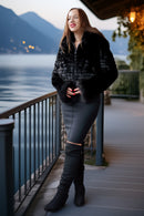 A woman stands on a lakeside boardwalk wearing a black faux-fur cropped jacket, a dark gray fitted skirt, and black over-the-knee boots, with mountains and water in the background.