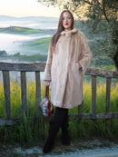 Woman in a beige coat standing in a scenic outdoor setting with a wooden fence and green fields.