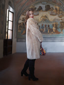 Woman in a long beige coat standing in a room with classical frescoes.