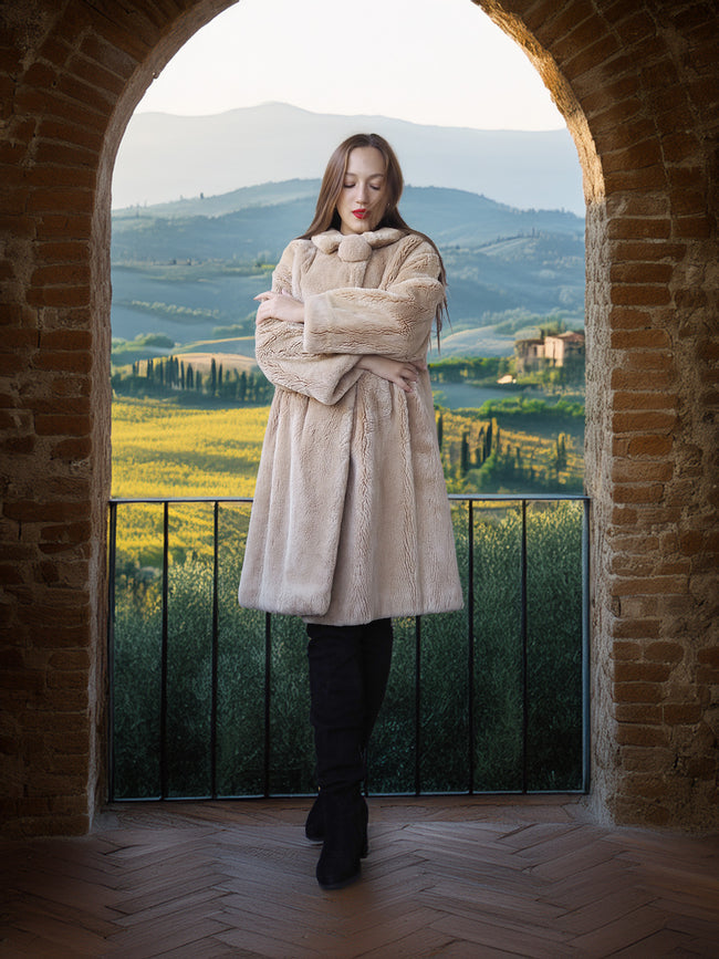 Woman in a beige coat standing in an archway with a scenic view of rolling hills and fields.