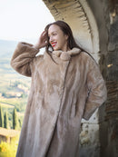 Woman wearing a fur coat standing against a stone wall with a scenic background