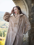 Woman wearing a fur coat standing against a stone wall with a scenic background