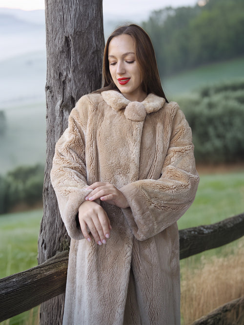 Woman wearing a beige fur coat standing next to a wooden fence with a scenic background.