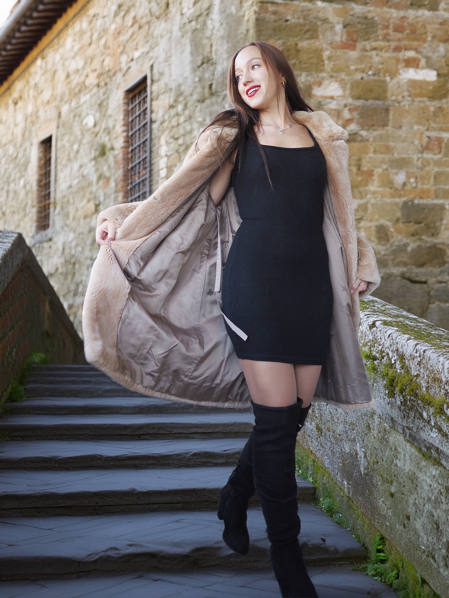 Woman in a black dress and knee-high boots standing on stone steps with a long coat over her shoulders.