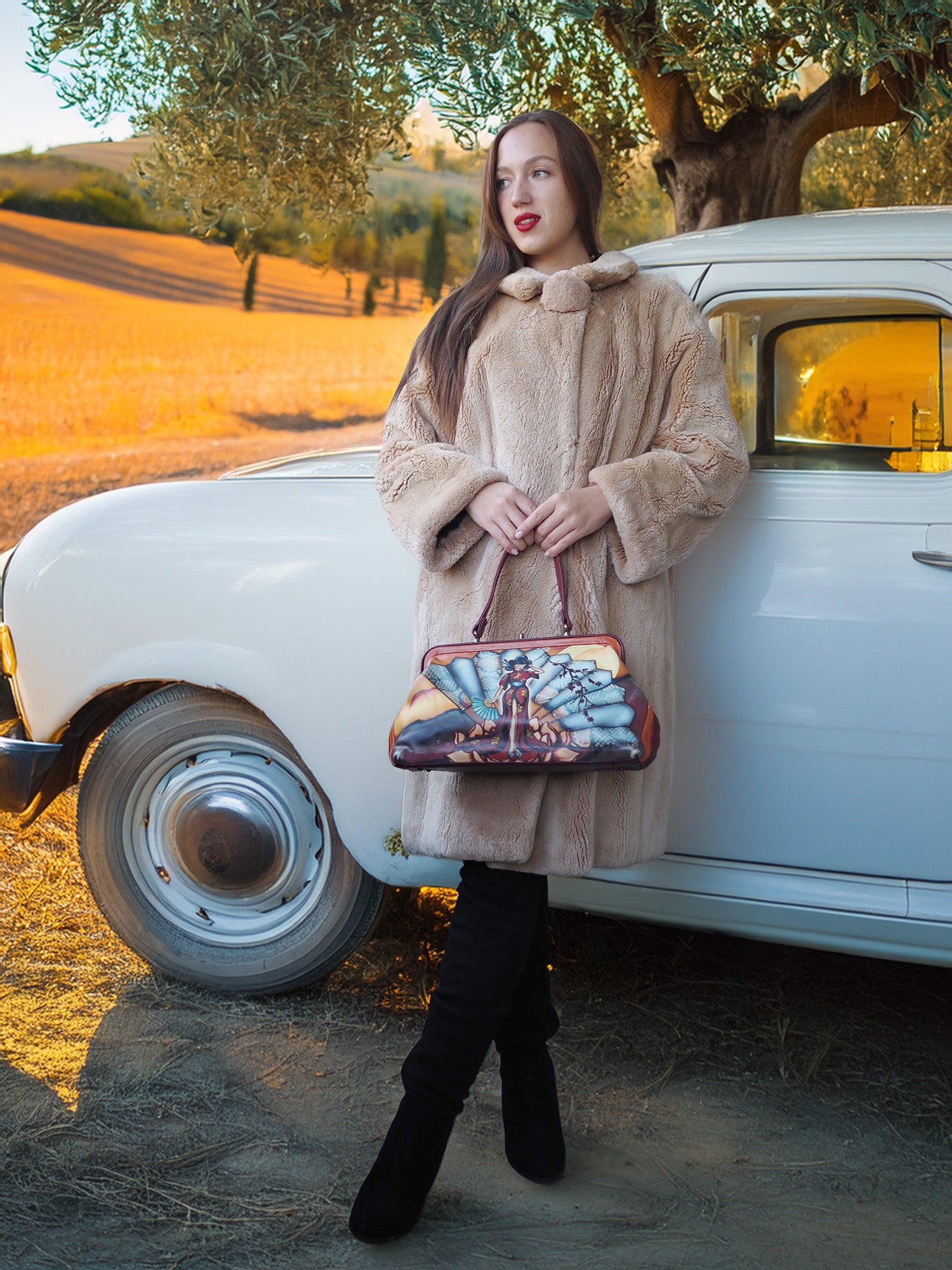 Woman in a beige coat holding a handbag with a scenic design, standing next to a vintage car in a rural setting.