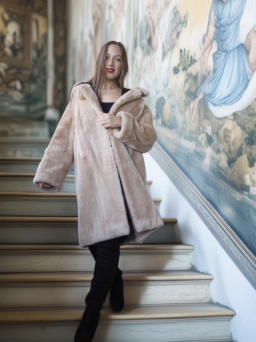 Woman in a beige fur coat standing on a staircase with decorative wall art in the background