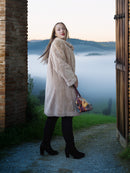 Woman in a beige coat standing in a scenic outdoor setting with a misty landscape.