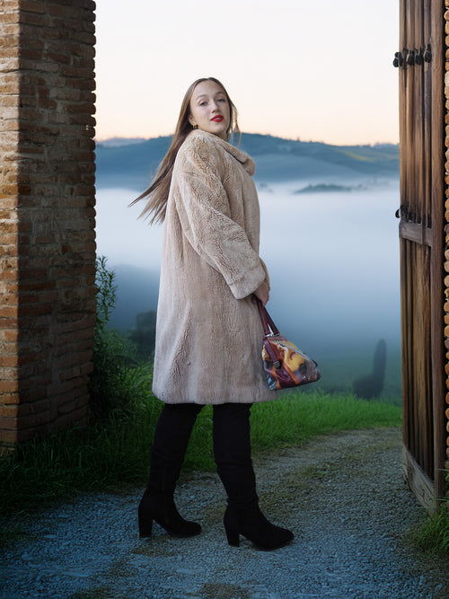 Woman in a beige coat standing in a scenic outdoor setting with a misty landscape.