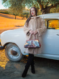 Woman in a beige coat holding a handbag with a scenic design, standing next to a vintage car in a rural setting.