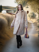 Woman in a long beige coat walking outdoors with a vintage car and trees in the background