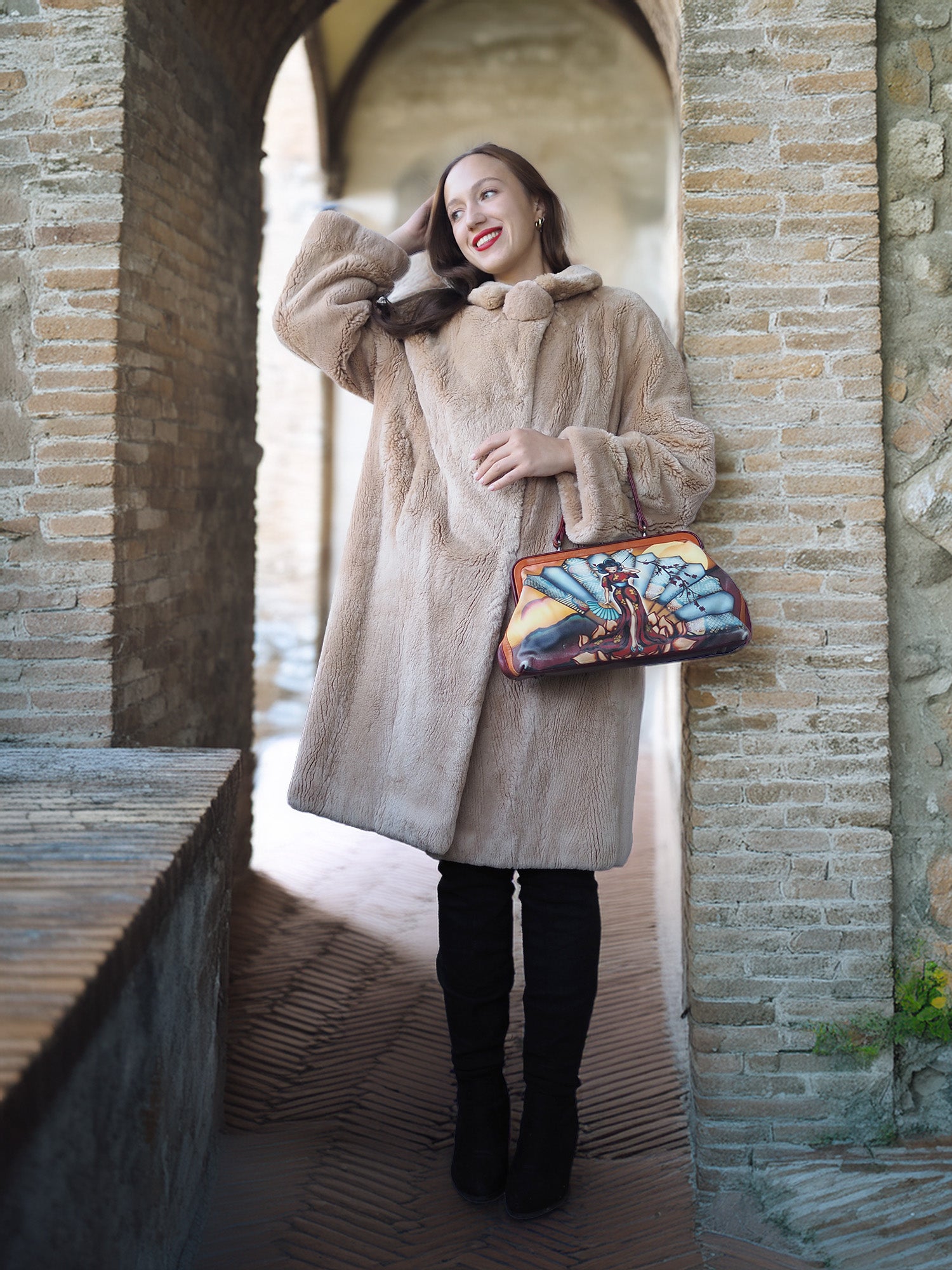 Woman in a beige coat holding a colorful handbag in a stone archway.