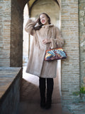 Woman in a beige coat holding a colorful handbag in a stone archway.