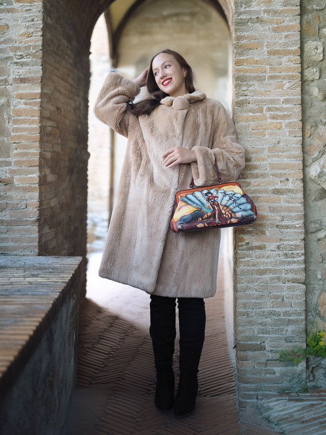 Woman in a beige coat holding a colorful handbag in a stone archway.