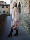 Woman in a beige coat holding a colorful handbag on a stone bridge.