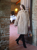 Woman in a beige coat standing in a rustic interior setting with brick walls and wooden door.