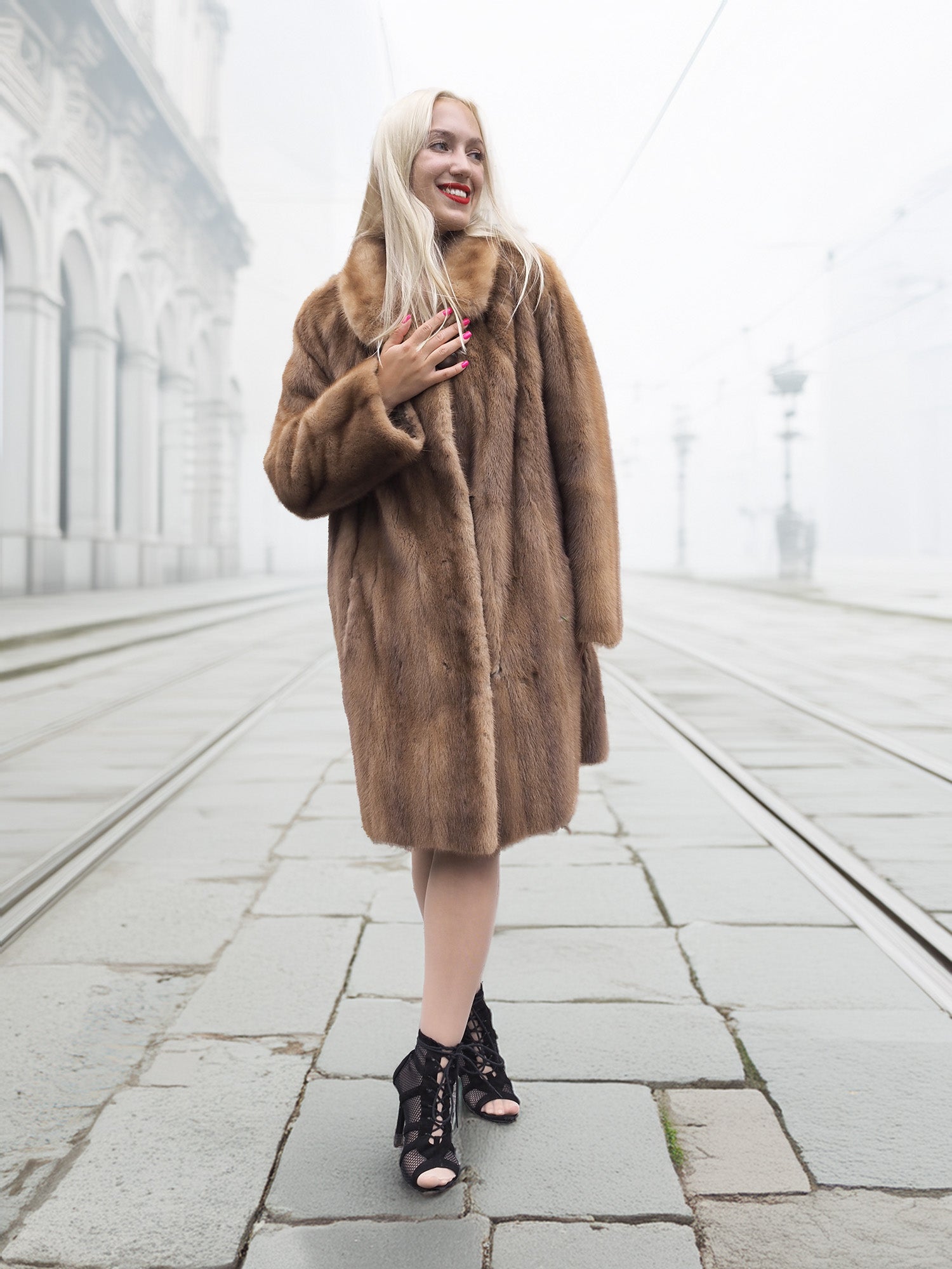 Smiling model in classic pastel brown mink fur coat stroller jacket M/L on foggy tramway. Luxurious and timeless fur fashion piece.