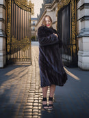 Woman in a fur coat standing in front of an ornate gate on a sunlit street.