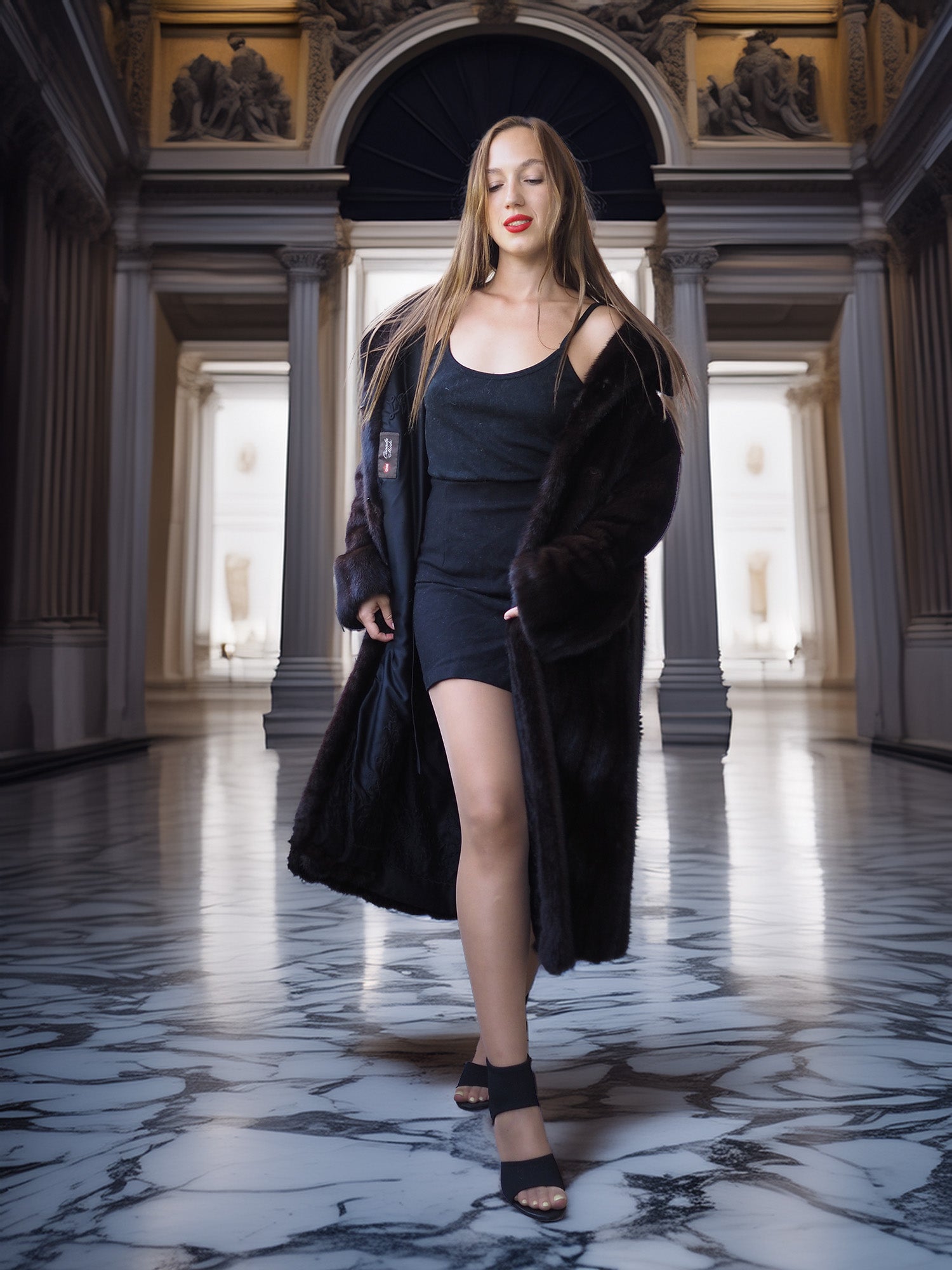 Woman in a long black coat standing in an elegant hallway with marble floors and classical architecture.