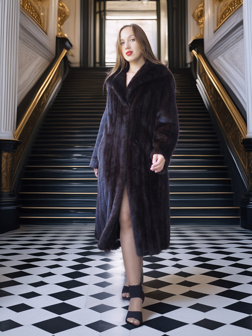 Woman in a long dark coat standing in an elegant interior setting with checkered floor and gold accents.