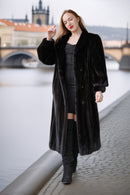 Woman in a long black coat standing by a waterfront with a bridge and buildings in the background.