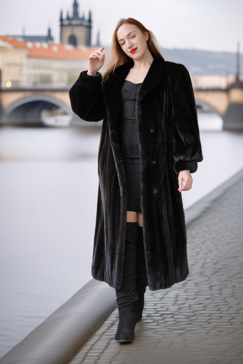 Woman in a long black coat standing by a waterfront with a bridge and buildings in the background.