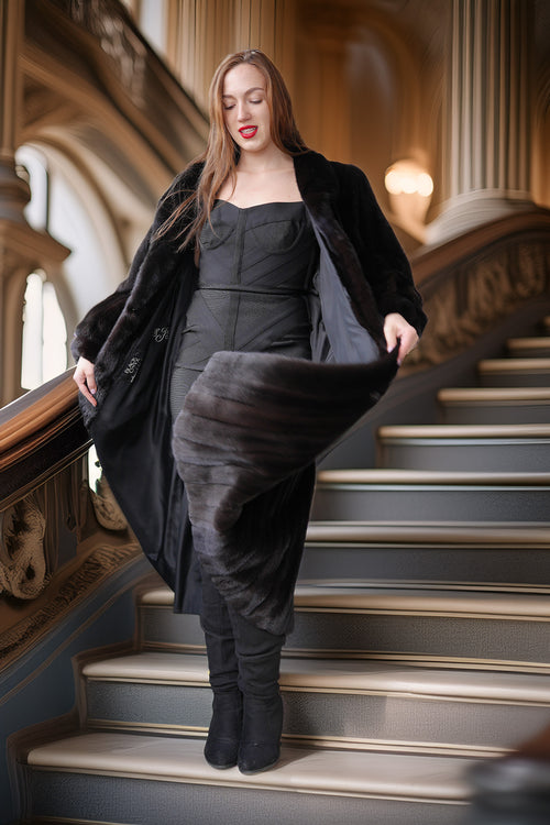 Woman in a black coat and dress standing on a staircase in an elegant interior setting