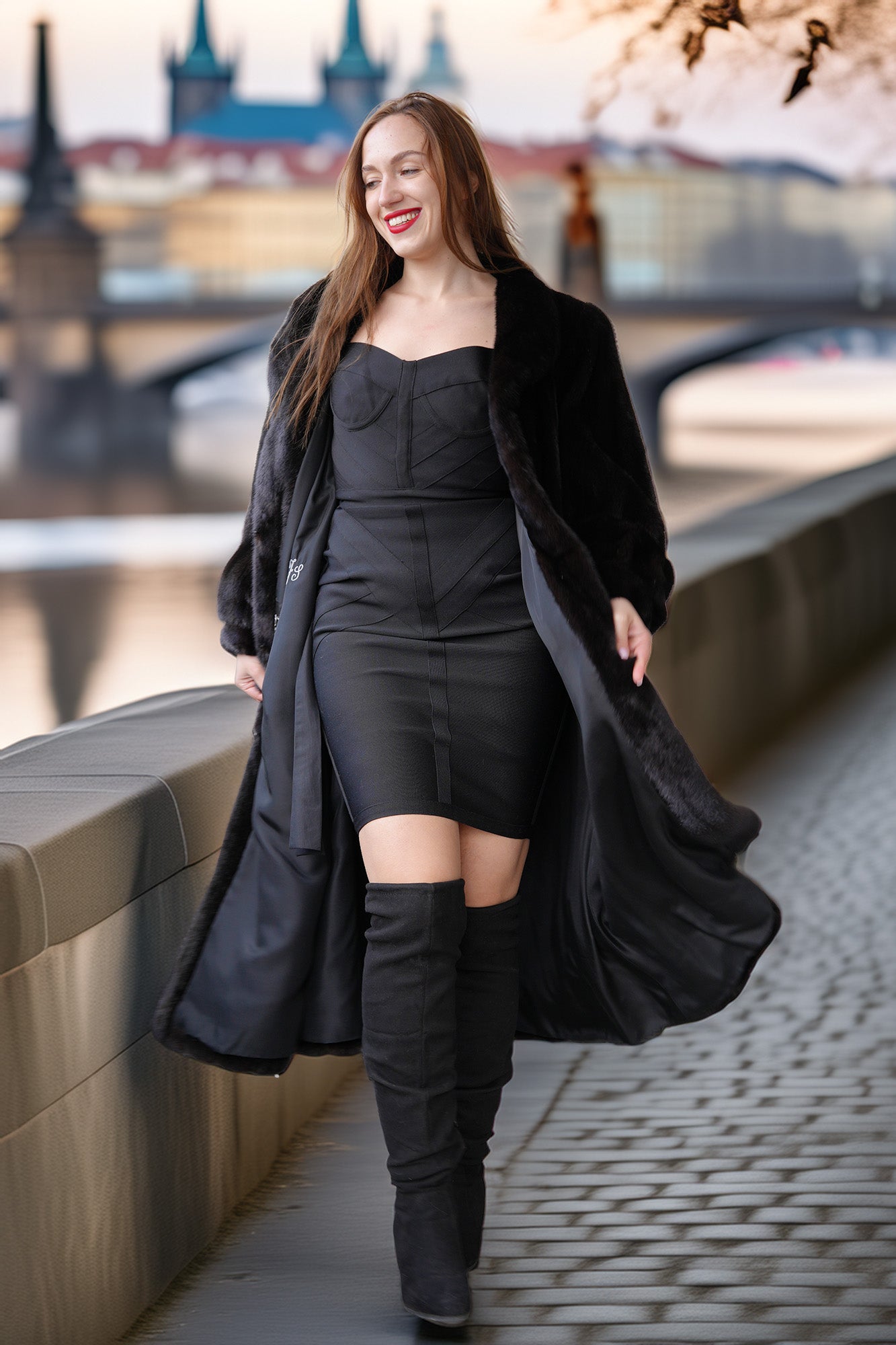 Woman in a black coat and dress with knee-high boots standing on a bridge with cherry blossoms in the background.
