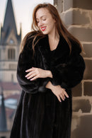 Woman wearing a black fur coat standing against a stone wall with architectural elements in the background.