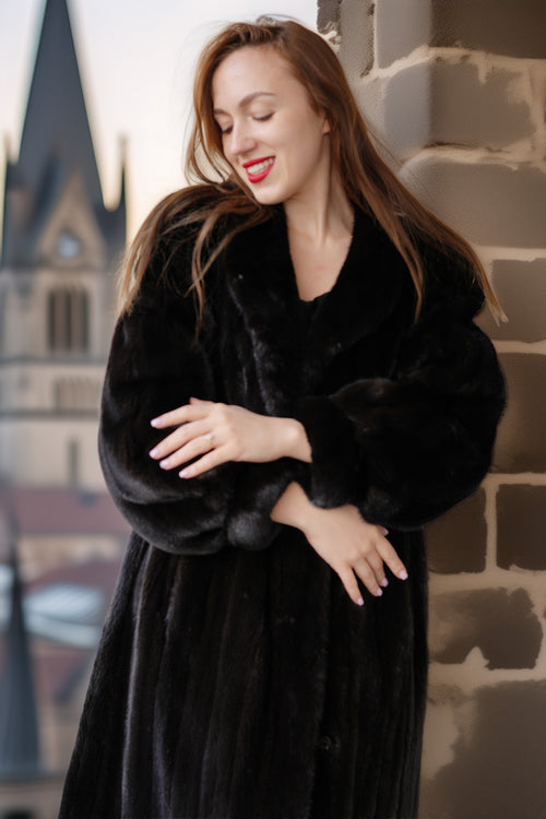 Woman wearing a black fur coat standing against a stone wall with architectural elements in the background.
