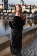Woman in a black fur coat standing by a river with a bridge and cityscape in the background