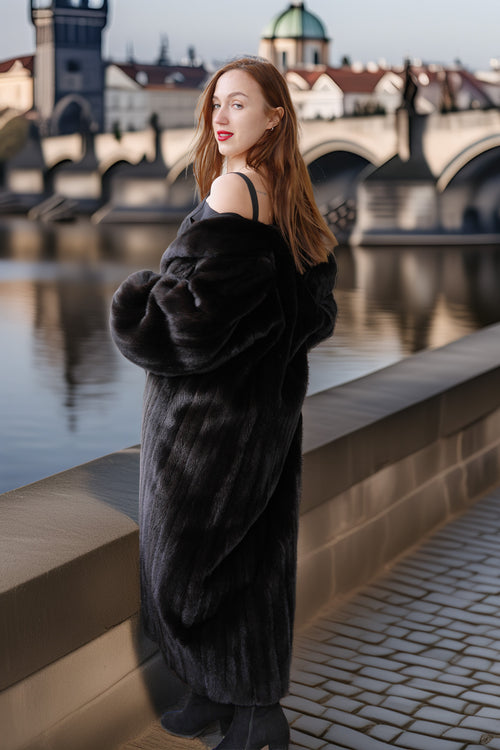 Woman in a black fur coat standing by a river with a bridge and cityscape in the background