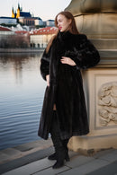Woman in a black fur coat standing by a waterfront with a cityscape in the background