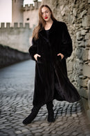 Woman in a black coat standing on a stone pathway with a castle-like structure in the background.