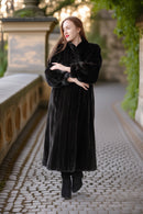 Woman in a long black coat walking on a cobblestone path with greenery in the background