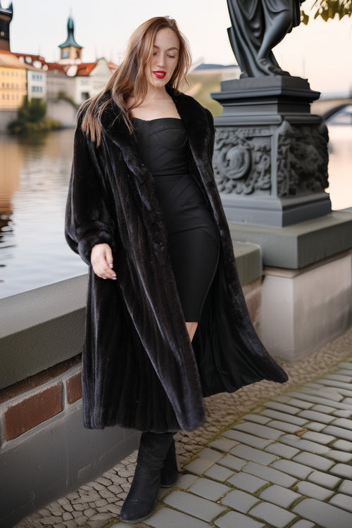 Woman in a long black coat standing by a waterfront with a scenic background