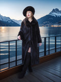 Woman in a long black coat and hat standing by a lake with mountains in the background