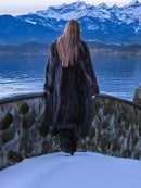 Person in a long coat standing by a snowy landscape with mountains and a lake.