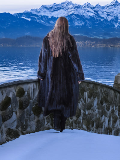 Person in a long coat standing by a snowy landscape with mountains and a lake.