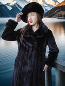 Woman in a fur coat and hat standing by a lake with mountains in the background