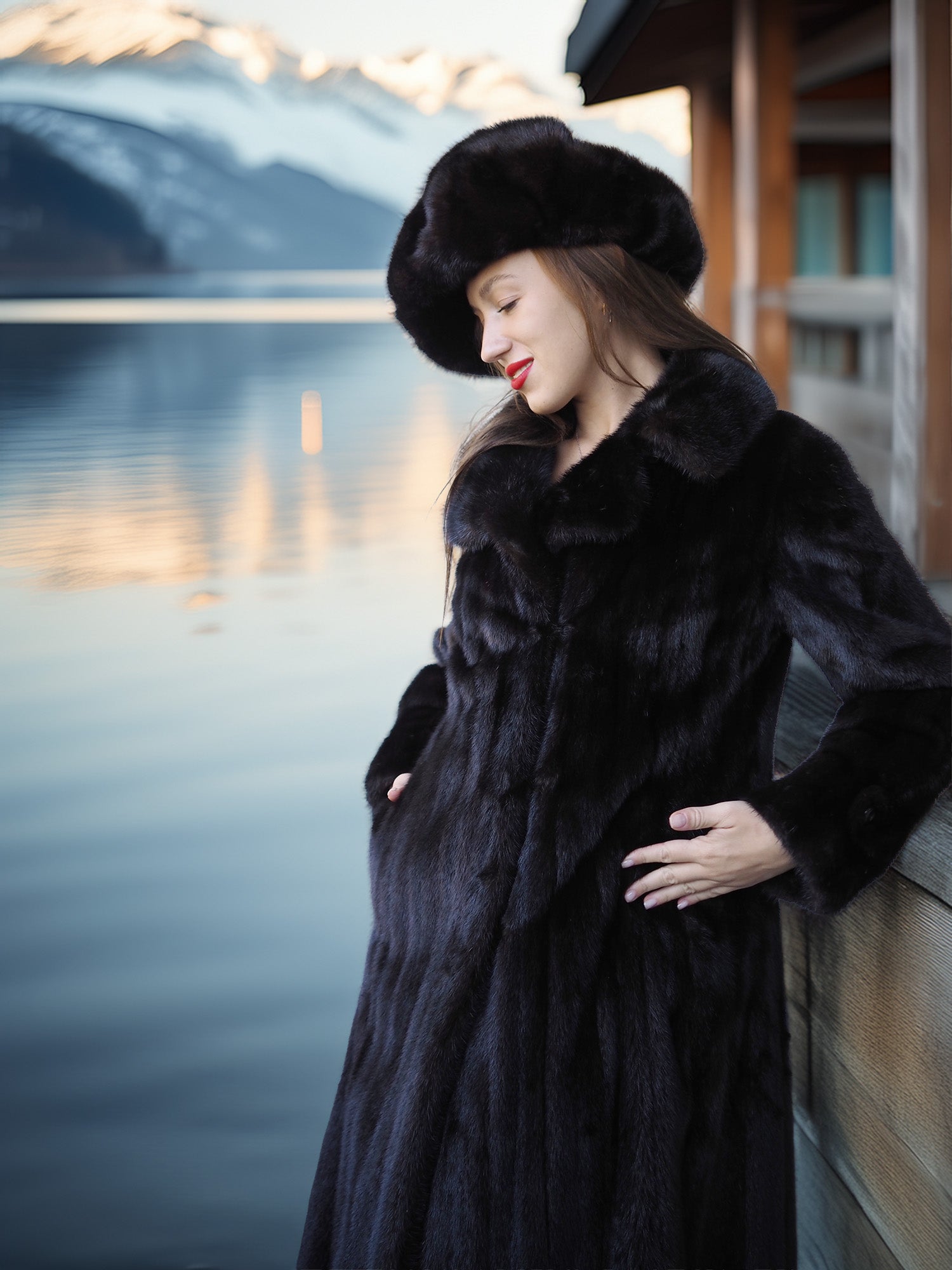 Woman in a black fur coat and hat standing by a body of water with mountains in the background.