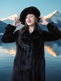 Woman in a black fur coat and hat standing in front of a mountainous landscape with water reflection.