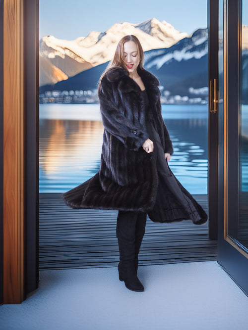 Woman in a long black fur coat standing in front of a large window with a mountain and lake view.