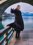 Person in a fur coat standing on a wooden dock with mountains and lake in the background
