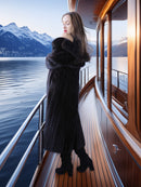 Woman in a long black coat standing on a boat with mountains in the background