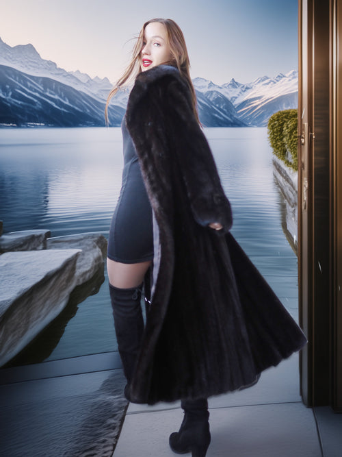 Woman in a long black fur coat standing by a scenic lake with mountains.