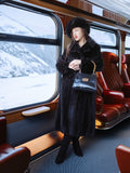 Woman in a black fur coat and hat standing on a train with snowy landscape outside.