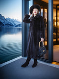 Woman in a fur coat and hat standing by a door with a scenic view of mountains and water.